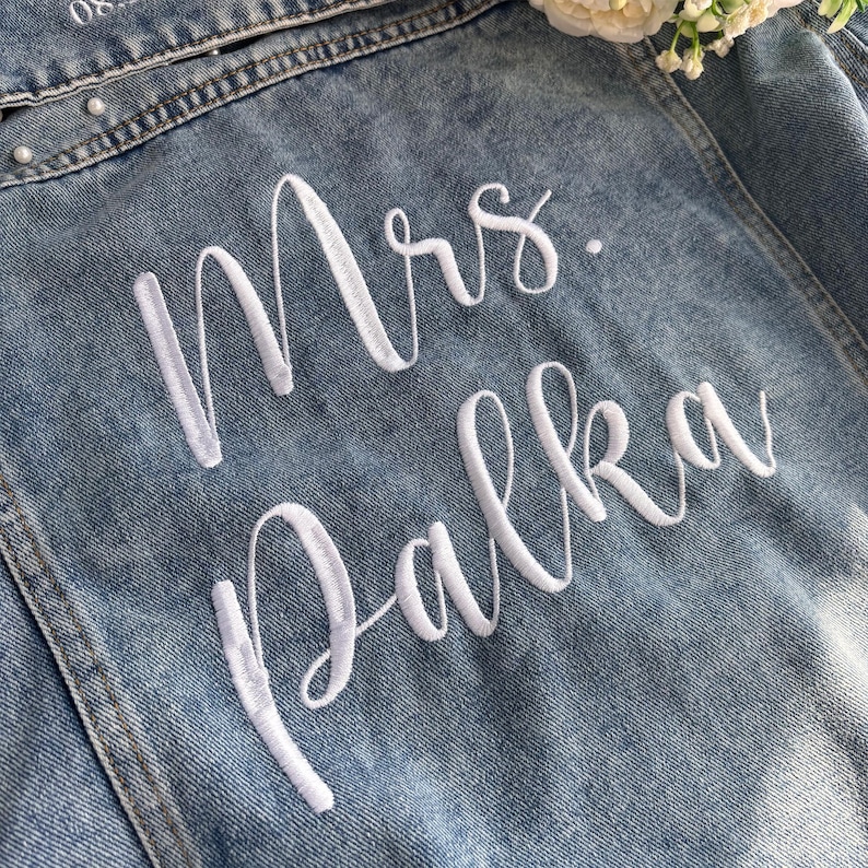 Puede incluir: Primer plano de una chaqueta vaquera azul claro con texto bordado en blanco. El texto dice "Mrs. Palka" en una fuente cursiva. La chaqueta tiene adornos de perlas a lo largo de la costura superior.