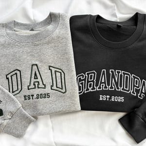 Peut inclure: Deux sweat-shirts gris et noirs avec les mots "DAD EST. 2025" et "GRANDPA EST. 2025" sur le devant. Les manches portent le nom "Cecilia Graham" et un cœur.