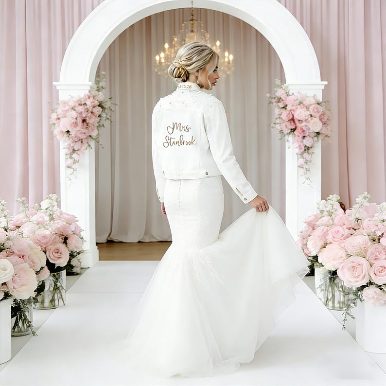 May include: A white denim jacket featuring the text Mrs. Stanbrook in cursive script on the back and a date on the collar, worn over a floor-length white mermaid-style gown with a flared tulle skirt and a fitted bodice with button details.