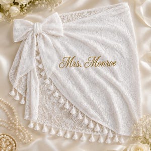 May include: A white lace sarong featuring a large decorative bow on the left side and a row of small white tassels along the hem. The fabric is embroidered with the gold text Mrs. Monroe in a cursive script, creating a detailed and elegant appearance.