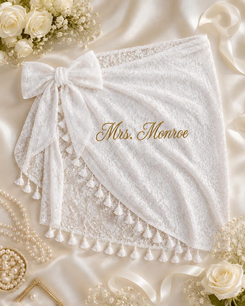 May include: A white lace sarong featuring a large decorative bow on the left side and a row of small white tassels along the hem. The fabric is embroidered with the gold text Mrs. Monroe in a cursive script, creating a detailed and elegant appearance.