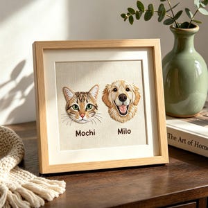 Może przedstawiać: Haftowane dzieło sztuki w drewnianej ramie przedstawiające kota i psa. Kot, o imieniu Mochi, ma zielone oczy. Pies, o imieniu Milo, to golden retriever. Grafika znajduje się na kremowym tle.