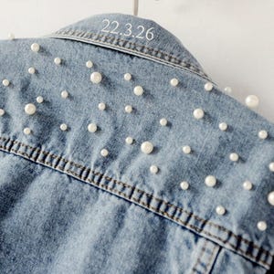 Puede incluir: Chaqueta vaquera azul claro adornada con perlas blancas dispersas. La chaqueta tiene un cuello cl&aacute;sico y una parte delantera abotonada. El texto "22.3.26" est&aacute; impreso en el cuello trasero. La chaqueta est&aacute; colgada en una percha blanca.