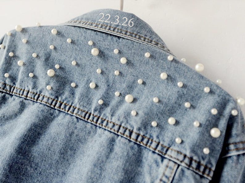 Puede incluir: Chaqueta vaquera azul claro adornada con perlas blancas dispersas. La chaqueta tiene un cuello cl&aacute;sico y una parte delantera abotonada. El texto "22.3.26" est&aacute; impreso en el cuello trasero. La chaqueta est&aacute; colgada en una percha blanca.