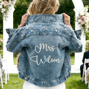 Puede incluir: Chaqueta vaquera azul claro con adornos de perlas y el texto bordado "Mrs. Wilson". La fecha "22.3.26" est&aacute; bordada en el cuello. La chaqueta se usa en una ceremonia de boda al aire libre.