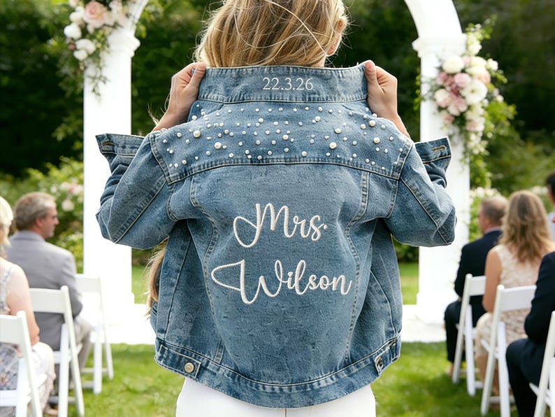Puede incluir: Chaqueta vaquera azul claro con adornos de perlas y el texto bordado "Mrs. Wilson". La fecha "22.3.26" est&aacute; bordada en el cuello. La chaqueta se usa en una ceremonia de boda al aire libre.