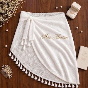 May include: A white lace wrap skirt featuring a side tie with hanging tassels and the gold embroidered text Mrs. Monroe. The skirt is decorated with a floral lace pattern and a scalloped hemline adorned with small white tassels along the edge.