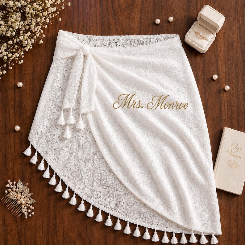 May include: A white lace wrap skirt featuring a side tie with hanging tassels and the gold embroidered text Mrs. Monroe. The skirt is decorated with a floral lace pattern and a scalloped hemline adorned with small white tassels along the edge.