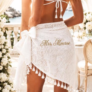 May include: A white lace sarong wrap featuring a side tie and a decorative tassel fringe along the hem. The fabric is embroidered with the text Mrs. Monroe in a gold script font. The garment is worn over a white bikini top with thin straps.