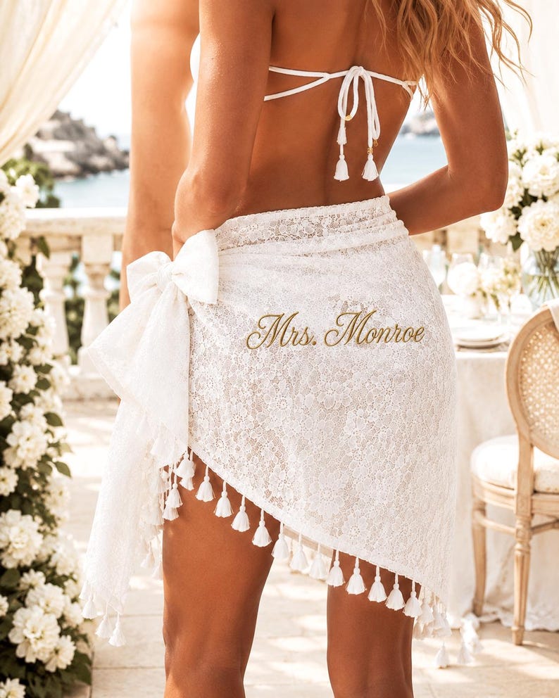 May include: A white lace sarong wrap featuring a side tie and a decorative tassel fringe along the hem. The fabric is embroidered with the text Mrs. Monroe in a gold script font. The garment is worn over a white bikini top with thin straps.
