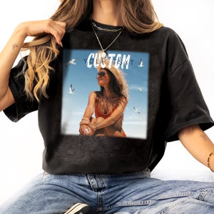 Może przedstawiać: Czarny T-shirt z nadrukiem graficznym przedstawiającym kobietę w bikini siedzącą na plaży z latającymi nad nią mewami. Na nadruku widnieje tekst "CUSTOM YOUR TEXT".