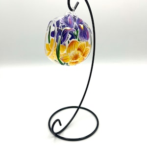 May include: A glass globe ornament with a painted floral design featuring yellow and purple flowers. The ornament is hanging from a black metal stand.