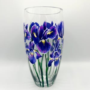 Peut inclure: Un vase en verre transparent avec un motif peint à la main de fleurs d'iris violettes. Le vase a un fond arrondi et une forme cylindrique.