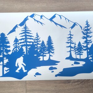 K&ouml;nnte beinhalten: Ein blauer Vinyl-Aufkleber mit der Silhouette einer Bigfoot-Figur, die durch eine Waldlandschaft mit hohen Kiefern, einem gewundenen Fluss mit verstreuten Steinen und einem gro&szlig;en Gebirgszug im Hintergrund auf einem wei&szlig;en rechteckigen Tr&auml;ger geht.