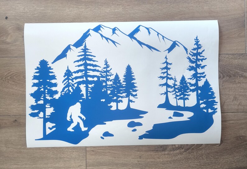 K&ouml;nnte beinhalten: Ein blauer Vinyl-Aufkleber mit der Silhouette einer Bigfoot-Figur, die durch eine Waldlandschaft mit hohen Kiefern, einem gewundenen Fluss mit verstreuten Steinen und einem gro&szlig;en Gebirgszug im Hintergrund auf einem wei&szlig;en rechteckigen Tr&auml;ger geht.