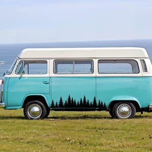 May include: A two-tone turquoise and white vintage camper van featuring a black silhouette decal of a pine tree forest along the lower side panel. The vehicle has chrome hubcaps, a high roof, and large windows, parked on a grassy field.