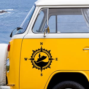 Peut inclure: Un van jaune présente un autocollant noir sur son panneau latéral. L'autocollant est un motif de rose des vents circulaire avec les lettres N, S, E et W aux points cardinaux. À l'intérieur, une silhouette de personne sur un jet ski est visible.