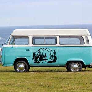 K&ouml;nnte beinhalten: Ein zweifarbiger t&uuml;rkiser und wei&szlig;er Vintage-Van mit einem schwarzen Vinyl-Aufkleber auf der Seitenwand. Der Aufkleber zeigt eine detaillierte Landschaftsszene mit einem Gebirge, einem dichten Kiefernwald und der Silhouette von Bigfoot.