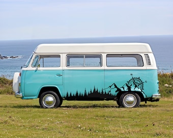 Könnte beinhalten: Ein zweifarbiger türkiser und weißer Vintage-Campingbus mit einem schwarzen Vinyl-Aufkleber auf der Seitenwand. Der Aufkleber zeigt die Silhouette eines dichten Kiefernwaldes, zerklüftete Berggipfel und eine nautische Kompassrose.