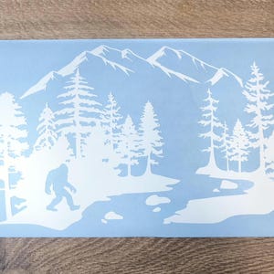 K&ouml;nnte beinhalten: Ein hellblauer rechteckiger Aufkleber mit einer wei&szlig;en Silhouettengrafik einer Bergkette, einem dichten Nadelwald, einem gewundenen Fluss und einer Bigfoot-Figur, die am Ufer entlanggeht, im Querformat.