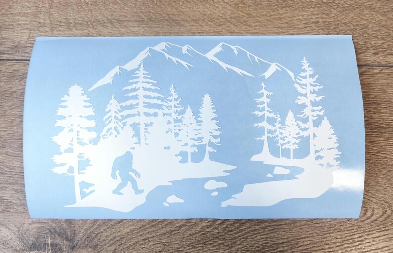 K&ouml;nnte beinhalten: Ein hellblauer rechteckiger Aufkleber mit einer wei&szlig;en Silhouettengrafik einer Bergkette, einem dichten Nadelwald, einem gewundenen Fluss und einer Bigfoot-Figur, die am Ufer entlanggeht, im Querformat.