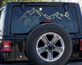 May include: A black Jeep SUV with a mountain and sun decal on the rear window. The decal is a yellow outline of a mountain range, a sun, and a tree. The spare tire is mounted on the back.