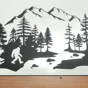 K&ouml;nnte beinhalten: Ein schwarzer Vinyl-Aufkleber mit der Silhouette einer Bigfoot-Kreatur, die durch einen Kiefernwald in der N&auml;he eines Flusses l&auml;uft, mit einer Bergkette im Hintergrund. Das Design ist auf einer wei&szlig;en Tr&auml;gerfolie dargestellt.