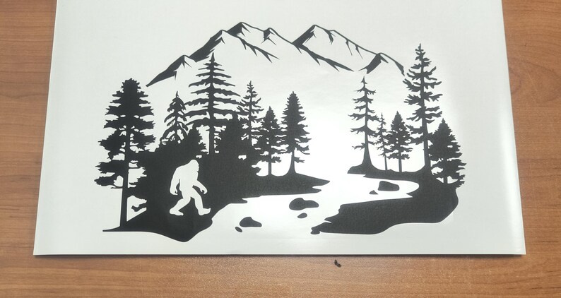K&ouml;nnte beinhalten: Ein schwarzer Vinyl-Aufkleber mit der Silhouette einer Bigfoot-Kreatur, die durch einen Kiefernwald in der N&auml;he eines Flusses l&auml;uft, mit einer Bergkette im Hintergrund. Das Design ist auf einer wei&szlig;en Tr&auml;gerfolie dargestellt.