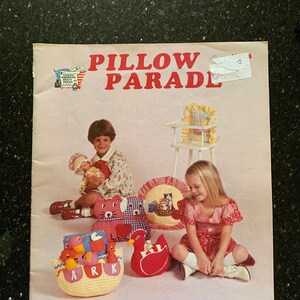 May include: A colorful book cover with the title "Pillow Parade" and the tagline "Pillows for Young and Old Alike". The cover features two children sitting on the floor surrounded by pillows shaped like animals, a boat, and a high chair.