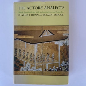May include: The cover of a book titled "The Actors' Analects" edited and translated by Charles J. Dunn and Bunzo Torigoe. The cover features a traditional Japanese painting of a procession of figures in a courtyard.