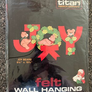 May include: A felt wall hanging kit featuring the word "JOY" with two teddy bears holding a wreath. The kit includes quality rayon/wool felt, trims, cotton floss, a needle, patterns, and complete illustrated instructions. The kit measures 21.6 cm by 49.5 cm.