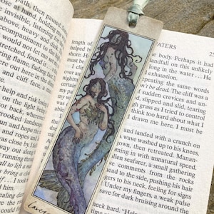 May include: A rectangular paper bookmark featuring a watercolor illustration of two mermaids with long dark hair and blue-toned tails. The bookmark has a light green ribbon tied through a hole at the top and the handwritten text Cavera Grace.