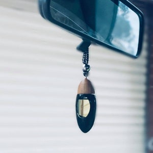 May include: A black car air freshener with a wooden top and a glass container. The air freshener is suspended from a rearview mirror. The liquid inside the container is visible.