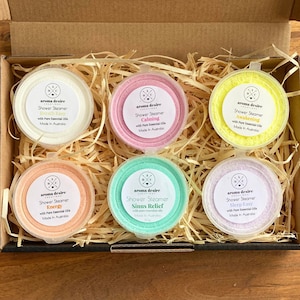 May include: A selection of six shower steamers in a gift box. The steamers are in shades of white, pink, yellow, orange, green, and purple. Each one is round and labelled with a different scent, including "Calming" and "Energy". The box is filled with straw.