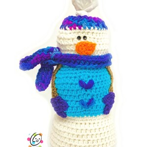 Può includere: Un giocattolo di pupazzo di neve all'uncinetto con un corpo bianco, una sciarpa blu e viola, un cappello blu con bordino bianco e un naso arancione.