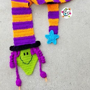 May include: A crocheted witch scarf with a purple and orange striped pattern. The scarf has a green witch face with a black hat and purple hair attached to one end. A blue star is attached to the other end.