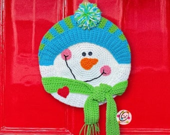 May include: A crocheted snowman wreath with a green scarf, pink cheeks, and a blue hat with a white pom-pom. The wreath is hanging on a red door.