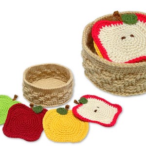 Puede incluir: Un juego de posavasos de ganchillo con forma de manzana en rojo, verde, amarillo y blanco. Los posavasos están en una cesta tejida con una manzana de ganchillo en su interior.