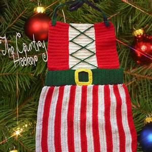 May include: Crocheted elf costume ornament with red, white, and green stripes. The ornament has a green belt with a yellow buckle and a green and white criss-cross pattern on the bodice. The ornament is hanging on a Christmas tree.