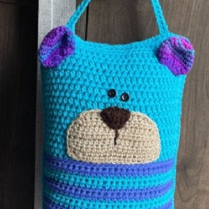 May include: A blue crocheted bear-shaped pillow with purple ears and a brown nose. The pillow has a striped pattern in blue and purple and is hanging from a door handle.
