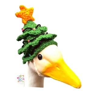 May include: A white goose wearing a green crocheted Christmas tree hat with a yellow star on top. The hat is decorated with black yarn and colorful lights. The goose has a yellow beak and a white neck.