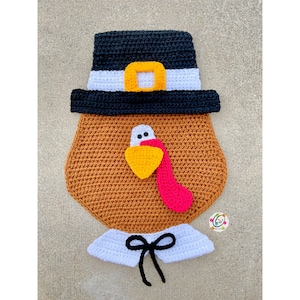 Può includere: Un tacchino all'uncinetto che indossa un cappello da pellegrino. Il tacchino è marrone con una bargiglio rosa e un becco giallo. Il cappello è nero con una fascia bianca e una fibbia bianca.