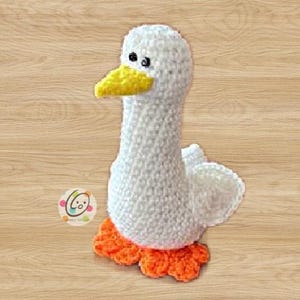 May include: A white crocheted goose with orange feet and a yellow beak. The goose has black eyes and is standing on a wooden surface.