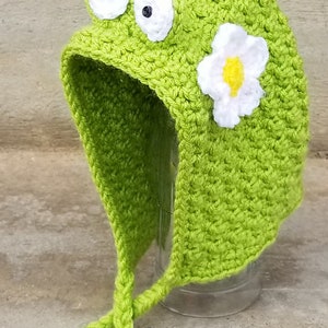 Könnte beinhalten: Grüne Häkelmütze mit einer Blume und zwei Glitz Augen. Die Mütze hat eine lange Schleife, die unter dem Kinn gebunden werden kann.