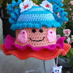 May include: A crocheted doll with a pink and orange dress, blue and white hat, and a white cat toy. The doll has a smiling face with pink cheeks and black eyes.