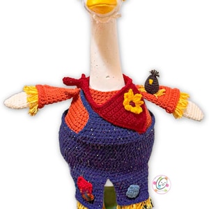 May include: A handcrafted goose ornament, dressed as a scarecrow. The goose is wearing a brown hat, orange and red crochet clothing, and blue overalls. It has yellow feet and a sunflower accent, ideal for a country-style home.