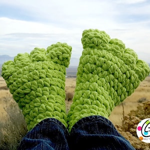May include: A pair of bright green, crocheted foot covers, resembling large feet, are visible against a natural outdoor backdrop. The textured, fuzzy material is a vibrant green. The person is wearing blue jeans. The logo for "scrappy-tots" is in the bottom right corner.