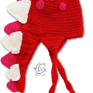 Könnte beinhalten: Rote und weiße gehäkelte Dinosauriermütze mit rosa Bommeln. Die Mütze hat einen langen Schwanz und eine herzförmige Nase.