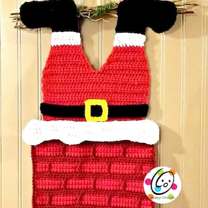 May include: A handmade Christmas decoration featuring Santa's legs and chimney, crocheted in red, black, white, and yellow. The chimney is brick-patterned, and the legs are upside down, with a black belt and yellow buckle. The decoration hangs from a twig base.
