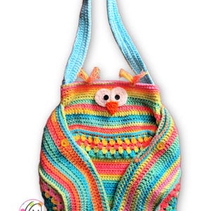 Może przedstawiać: Kolorowa, ręcznie robiona torba z sową zrobiona na szydełku z jasnoniebieskim paskiem. Torba ma tęczowe kolory, w tym różowy, pomarańczowy, żółty i niebieski. Twarz sowy ma duże białe oczy, pomarańczowy dziób i małe pomarańczowe akcenty.